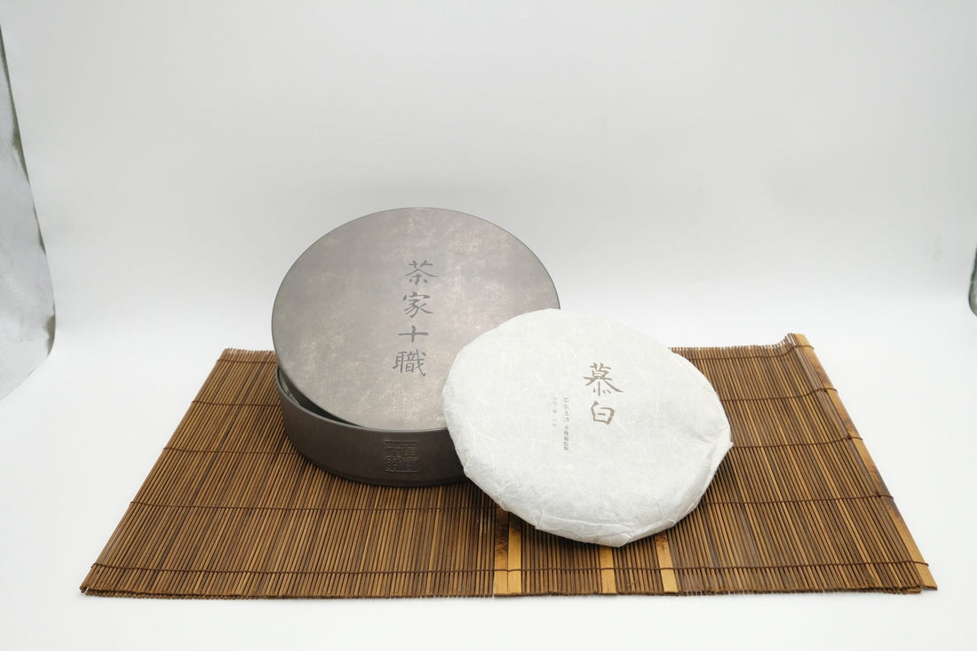 A compressed white tea cake in its paper wrapper on a bamboo mat beside a dark metal storage tin